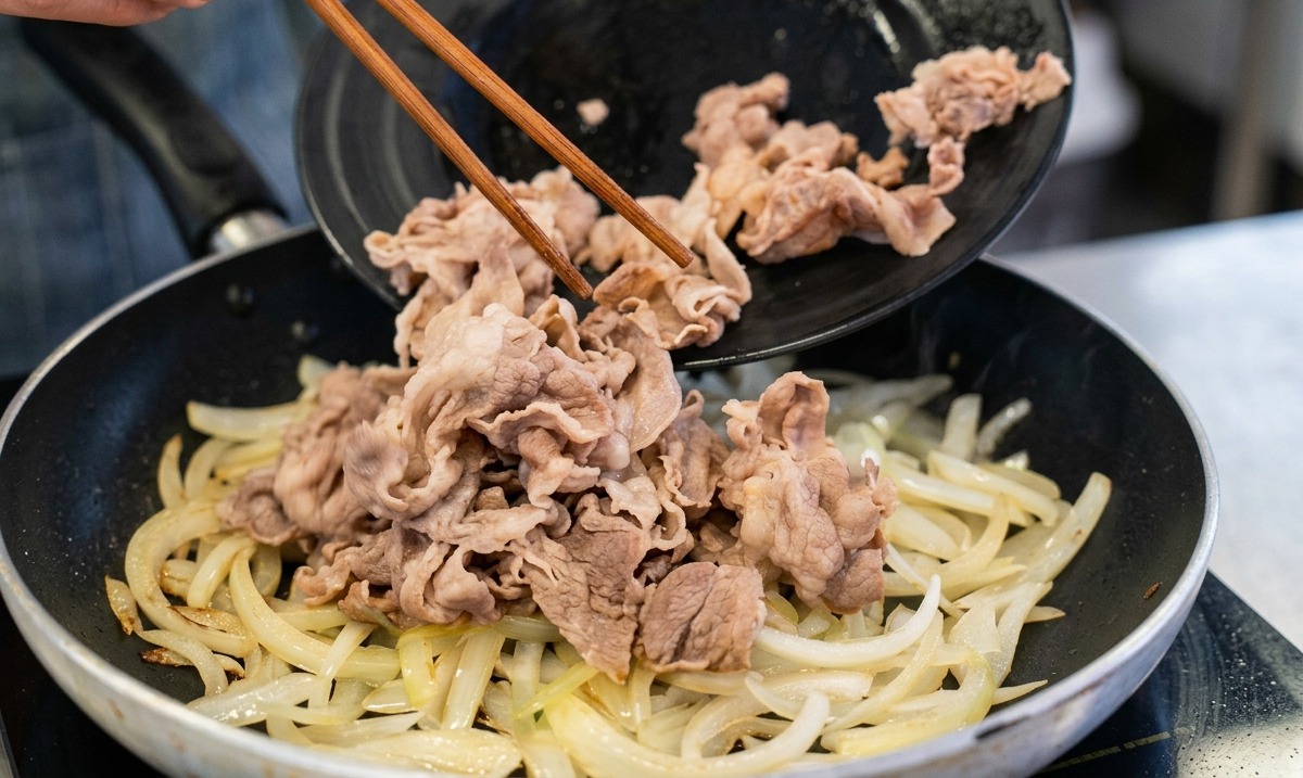 木箸を使い、黒いお皿に置いてあった下茹で済みの牛肉を、玉ねぎが炒められているフライパンへと滑らせるように入れている様子。