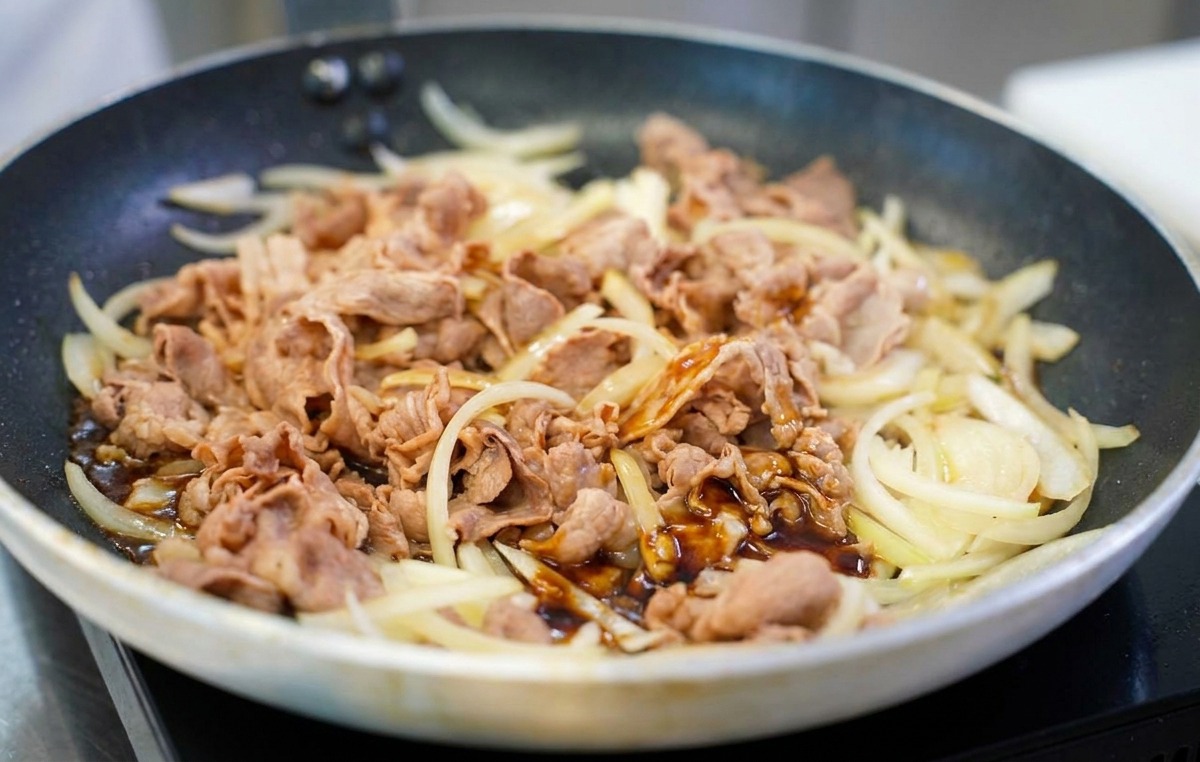 フライパンの中で炒められている牛肉と玉ねぎの上から、艶のある濃い茶色の合わせタレを均一に回しかけている様子。