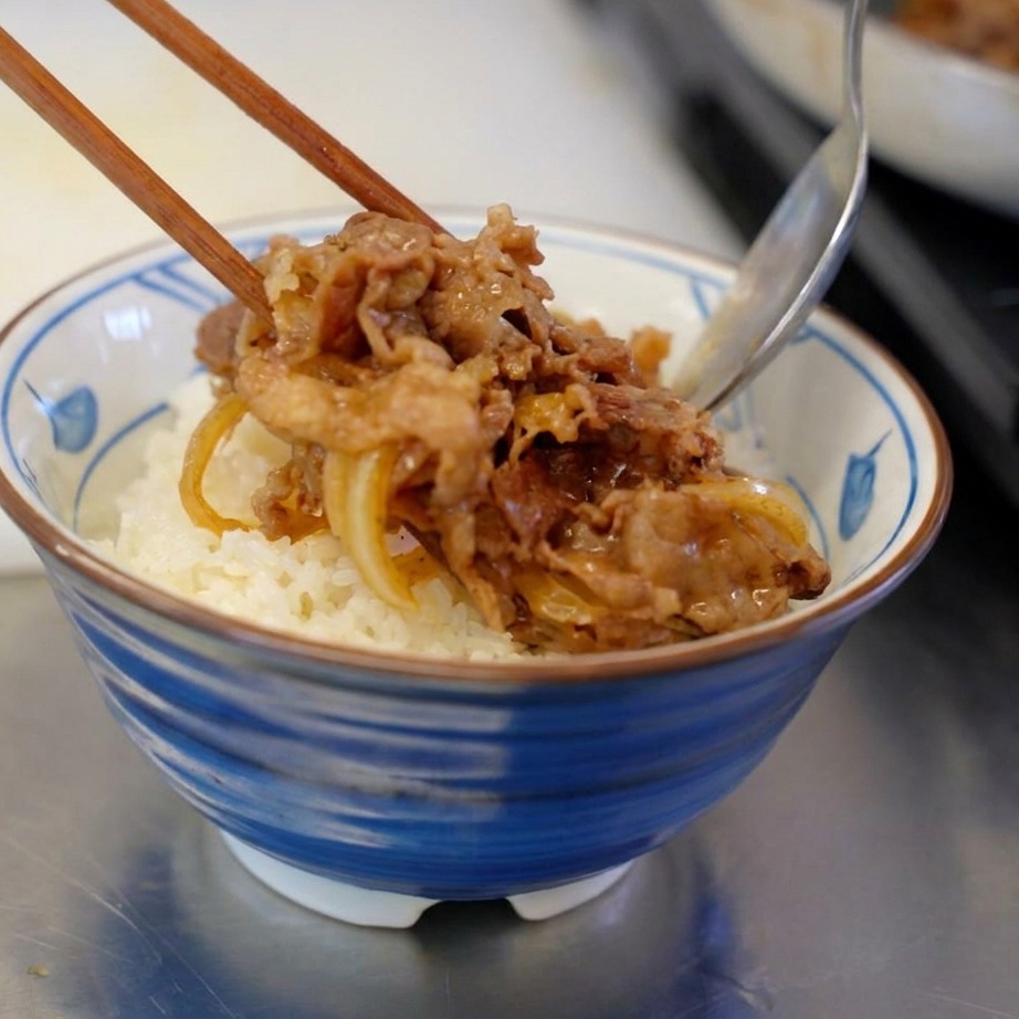 ほかほかの白いご飯が盛られた丼の上に、お箸とスプーンを使って、味がしっかり染みた牛肉と玉ねぎをたっぷりのせている様子。