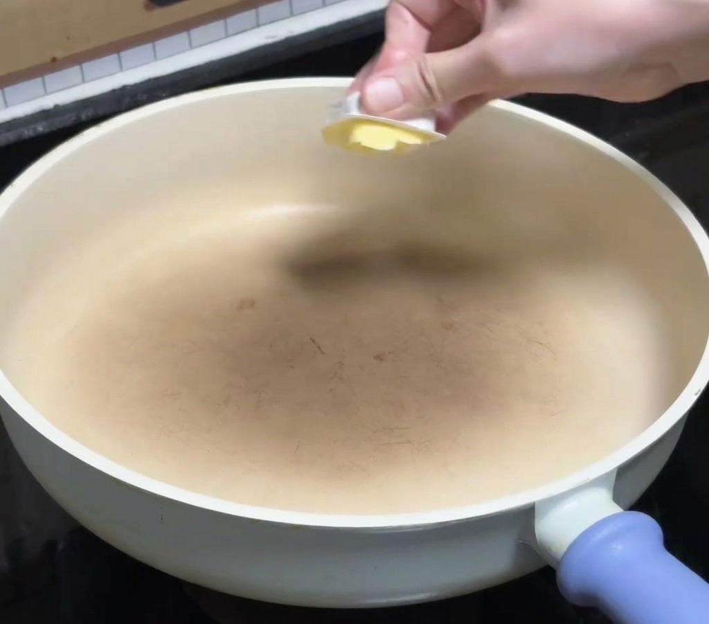 コンロの上の白いフライパンに、四角い黄色いバターを入れて溶かしている手元。
