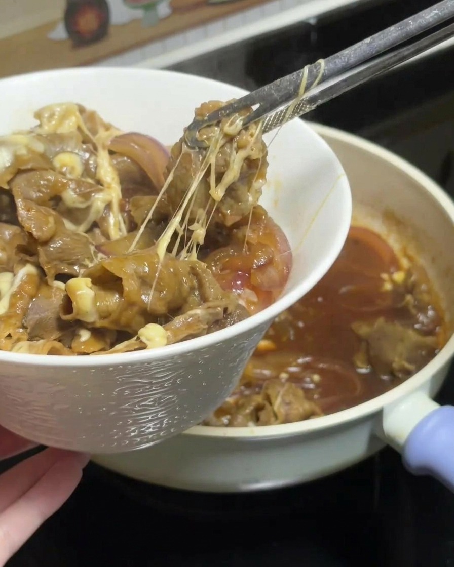 トングを使って、チーズが長く伸びた状態の照り焼きチキンを丼のご飯の上にのせている様子。
