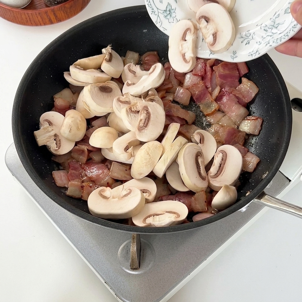 焼いたベーコンと玉ねぎの中に、スライスしたマッシュルームを入れる様子。