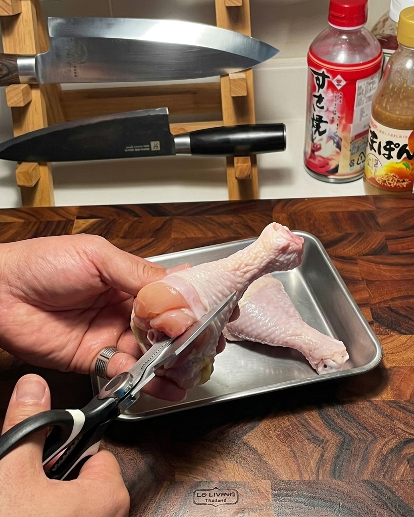 木製のまな板の上で、キッチンバサミを使って生の鶏もも肉から骨を丁寧に切り離している様子。