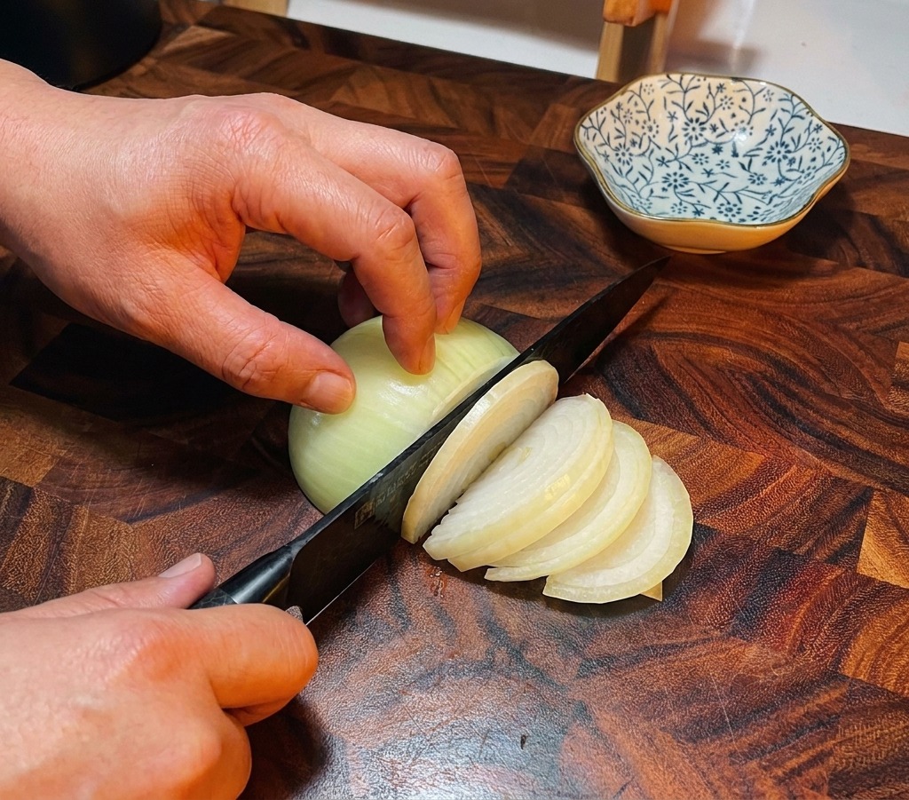 濃い色の木製まな板の上で、シェフナイフを使って玉ねぎを薄くスライスしている様子。