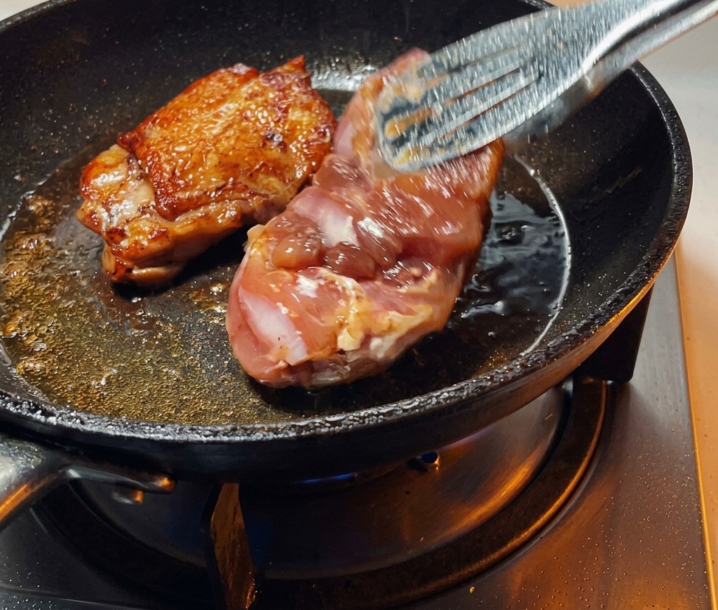トングを使ってフライパンの中の鶏もも肉をひっくり返し、完璧にこんがりと焼けた皮目を上に向けている様子。