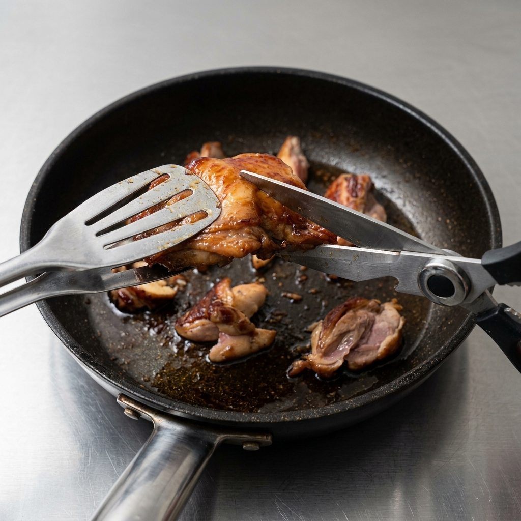 フライパンの中で、キッチンバサミを使ってこんがり焼けた鶏もも肉を小さめの一口大にチョキチョキと切っている様子。