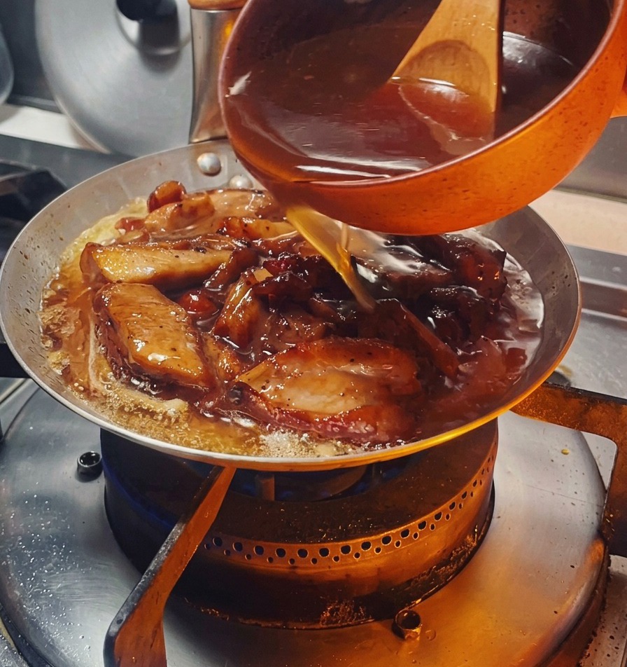 オレンジ色の小さなボウルから、こんがり焼けた鶏肉と玉ねぎが入ったフライパンへ、濃い色の甘辛いタレを回し入れている様子。