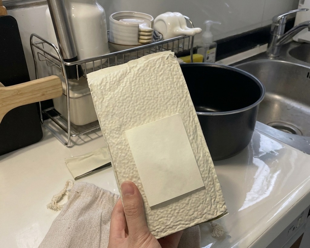 清潔なキッチンのカウンターの上で、白い布製の袋を持ち、炊飯器の黒い内釜に白い短粒米の乾燥米を注いでいる手。