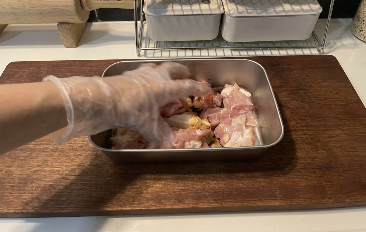 木製のまな板の上の浅い金属製トレイの中で、生の鶏肉の切れ端に濃い醤油ベースのマリネ液を混ぜ合わせている、透明なプラスチック手袋をはめた手。
