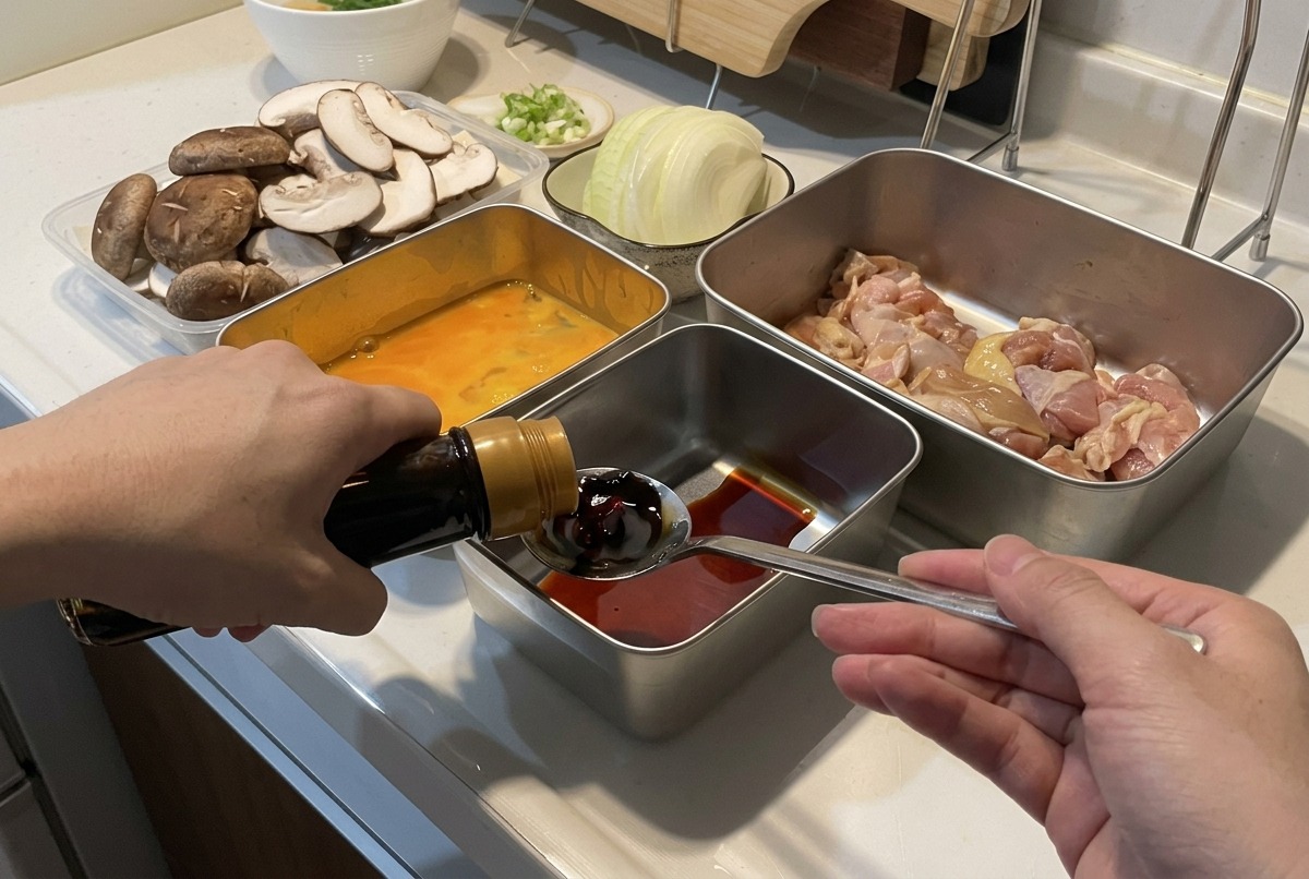 スライスした玉ねぎやキノコなどの下ごしらえされた食材に囲まれ、液体が入った金属容器の上のスプーンに、ボトルから濃い醤油を注いでいる人。