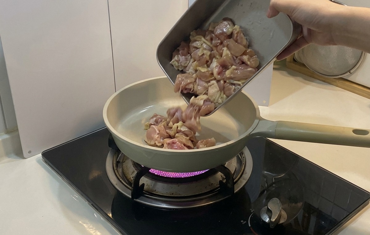 紫色のガス炎の上にある淡い色のセラミックフライパンに、下味をつけた鶏肉の切れ端を金属製トレイから注ぎ入れている様子。