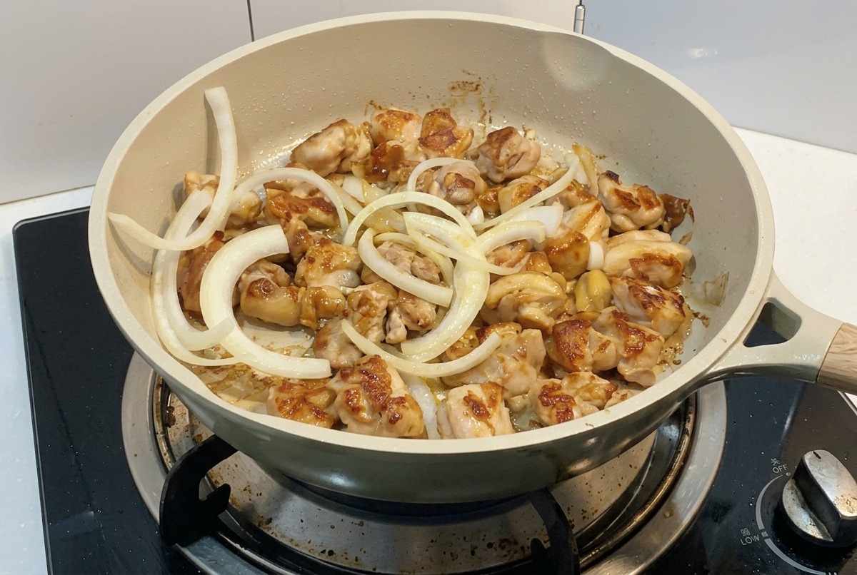 コンロの上の、焼き色がついてきた鶏肉の入ったフライパンに、スライスした白玉ねぎのリングを加えている様子。