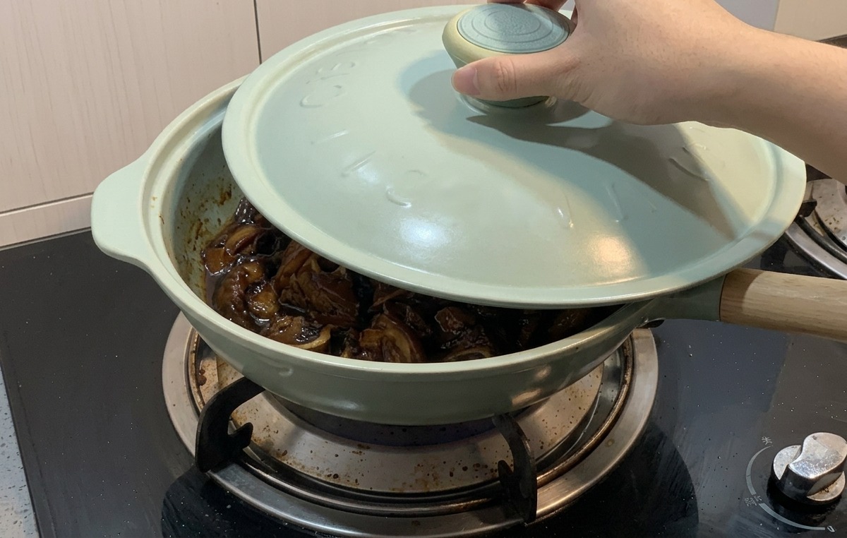 濃い色のタレで煮込まれている鶏肉と玉ねぎが入った薄緑色のフライパンに、手で薄青色の蓋をかぶせている様子。