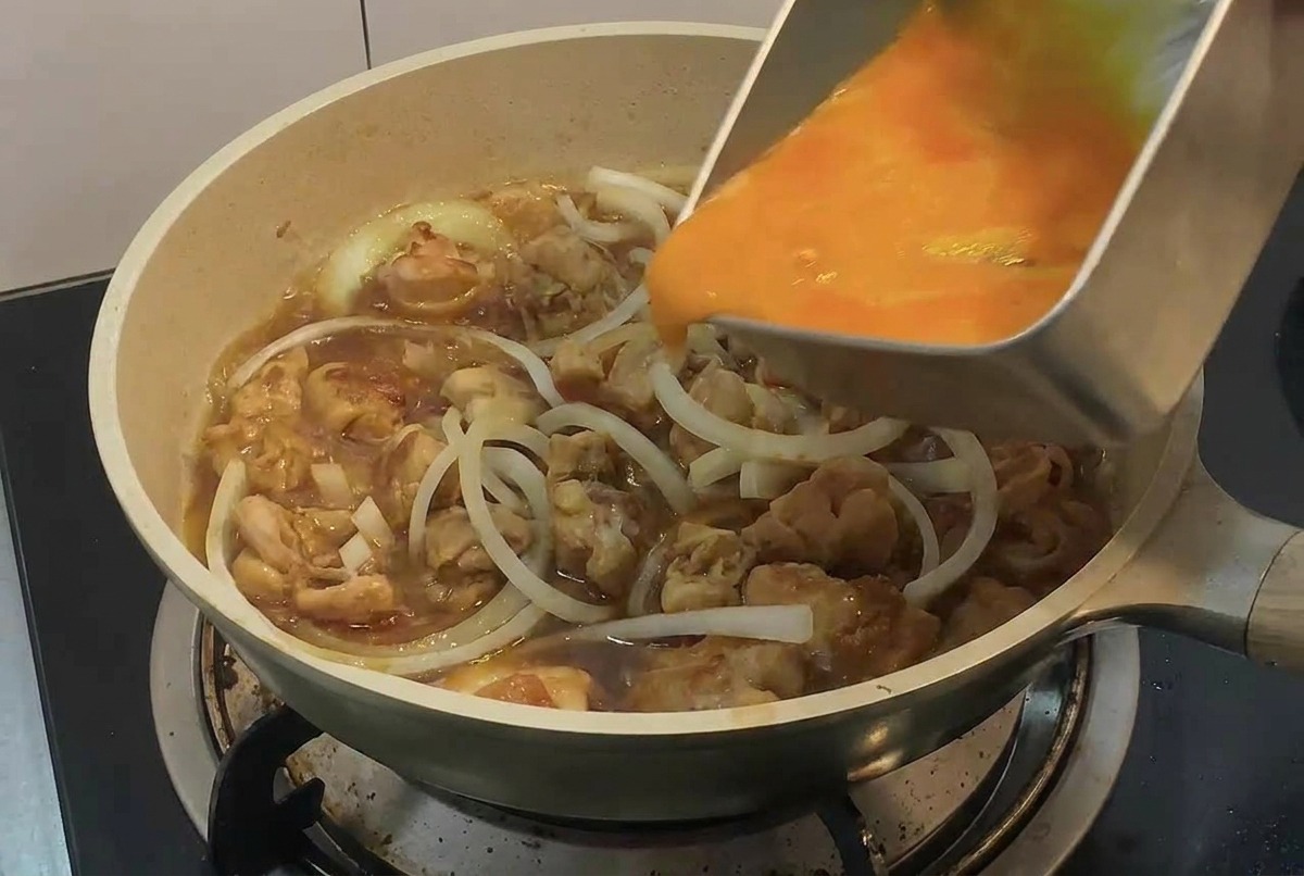 長方形の金属トレイから、鶏肉と玉ねぎのスライスが煮立っているフライパンに、粗く混ぜた卵液を注ぎ入れている手。