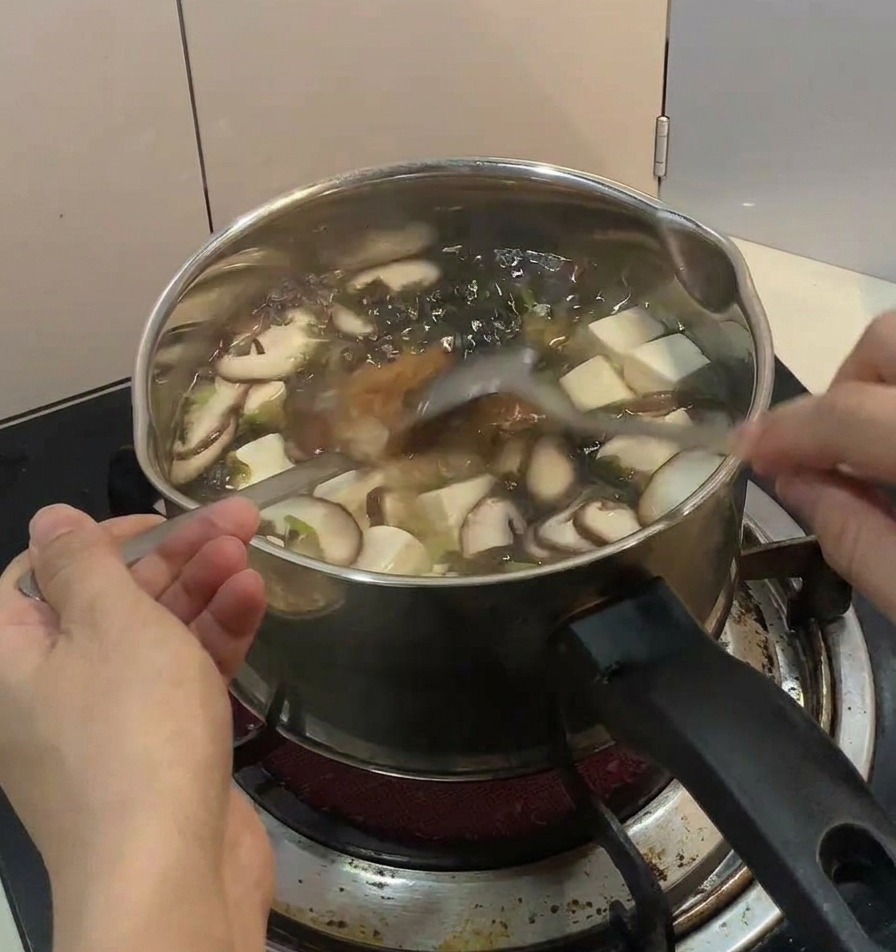木製のテーブルの上で、湯気が立つ鶏肉と卵の丼の上に、手で新鮮な青ネギのスライスと濃い色の海苔の細切りを散らしている様子。