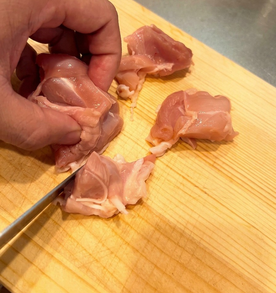 木製のまな板の上で、生の鶏もも肉を一口大の大きさに均一に切っている様子。