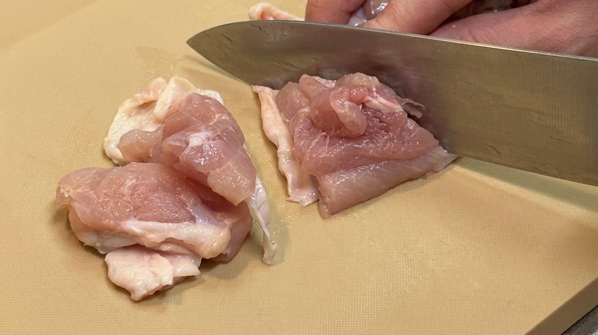 大きめの包丁を使って、生の鶏もも肉を一口大の大きさに均等に切り分けている様子。