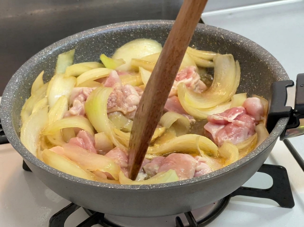 フライパンの中で、煮立つ濃い液体とともに半分火が通った鶏肉と玉ねぎを、木べらで優しく混ぜている様子。