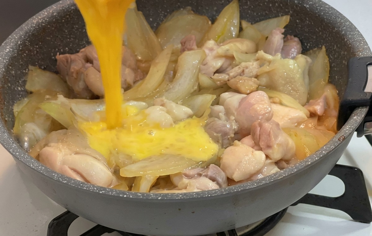 出汁で煮込まれる鶏肉と玉ねぎの上へ、小さなボウルから溶き卵を回し入れている様子。