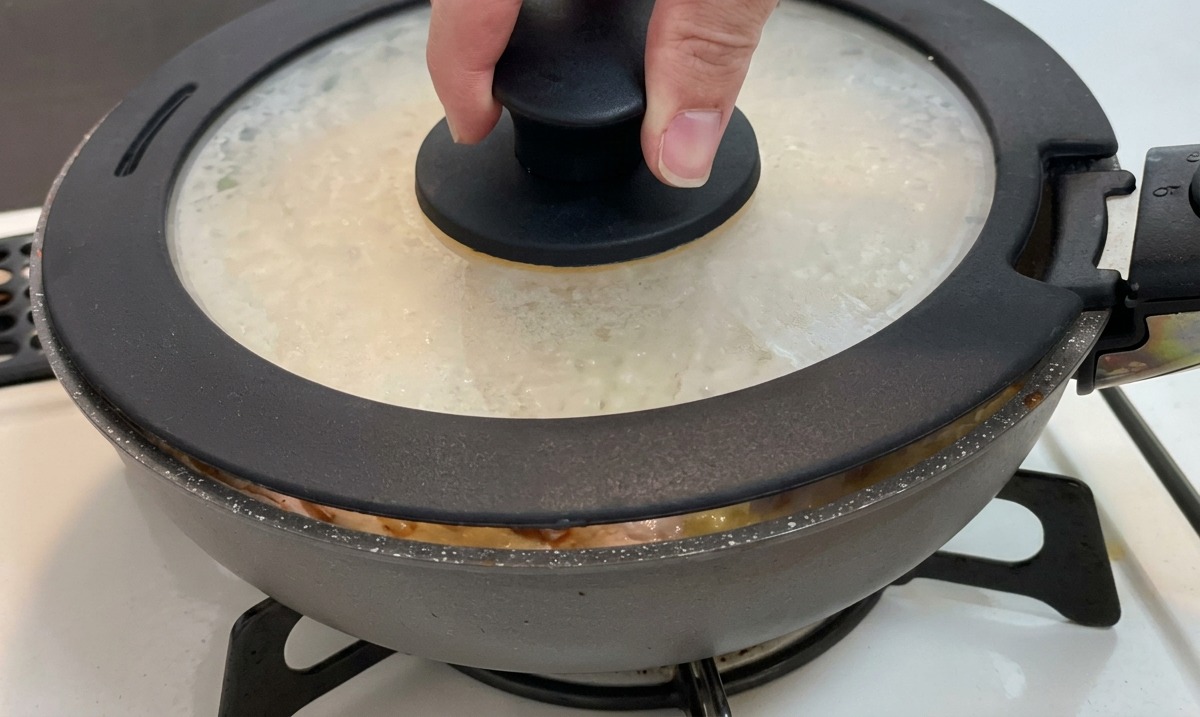 鶏肉、玉ねぎ、卵が入ったフライパンに、黒いツマミのついたガラス製の蓋をかぶせている手の様子。