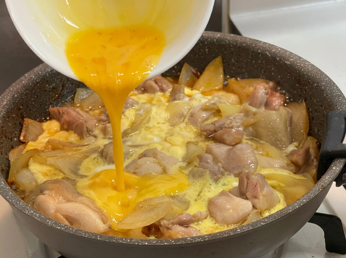 煮立つ鶏肉、玉ねぎ、半熟の卵が入った熱いフライパンへ、白いボウルから鮮やかな黄色の溶き卵を注いでいる様子。