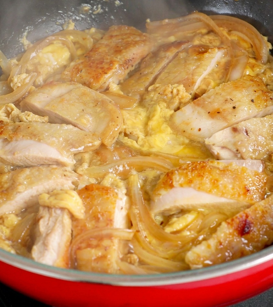 赤いフライパンの中で玉ねぎと馴染んだ鶏肉の上から、とろりとした卵液の残りを注き、極上の半熟仕立てにしている様子