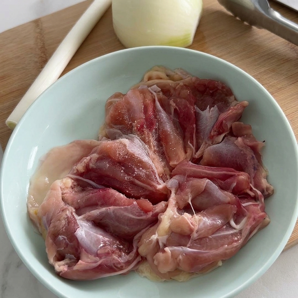 淡いブルーのボウルに入った生の鶏もも肉に、塩と挽きたての黒こしょうを振って下味をつけている様子。