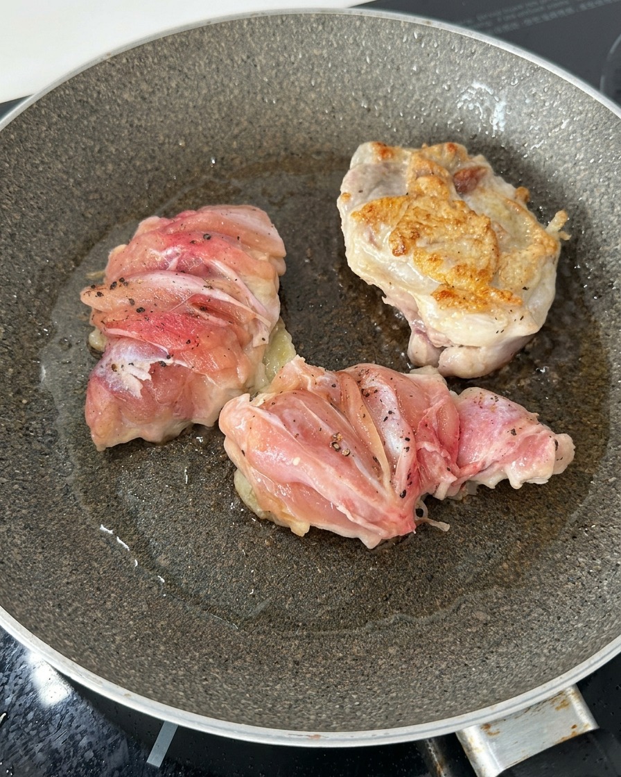 フライパンの中にある3つの鶏もも肉のうち、1つをひっくり返して、表面がこんがりと黄金色に焼けているのを見せている様子。