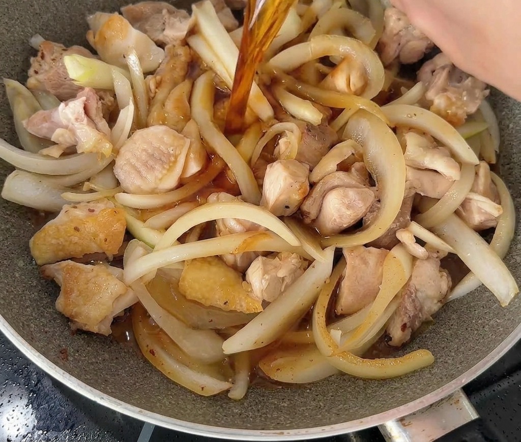 鶏肉と玉ねぎのスライスが入ったフライパンに、白いボウルから濃い色をした特製のタレを注いでいる様子。