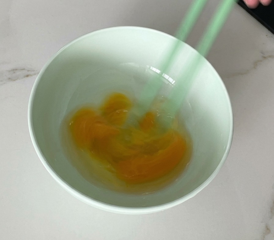 薄緑色のボウルの中で、緑色の箸を使って生の卵を軽く混ぜ、黄身と白身がマーブル状に混ざり合っている様子。
