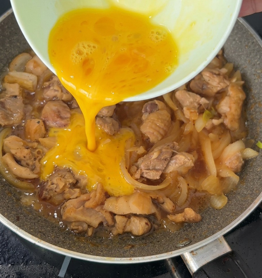 フライパンの中でグツグツと煮立つ鶏肉と玉ねぎの上に、溶き卵が細く注がれている様子。