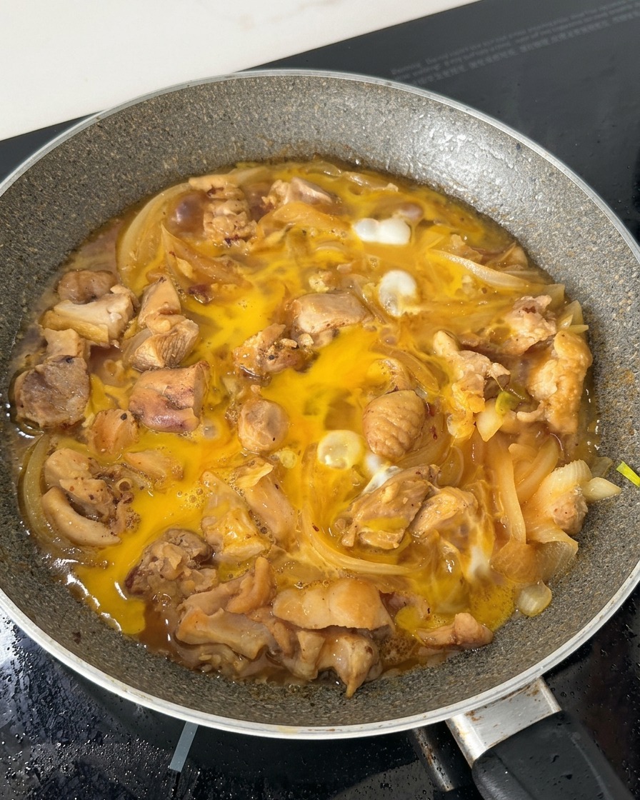 しょうゆベースの出汁の中で、鶏肉や玉ねぎと共に、半分固まりかけた卵液がフライパンの中でフツフツと泡立っている様子。