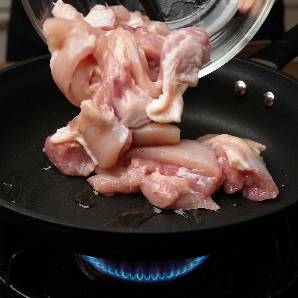 ガラスボウルから生の鶏肉のぶつ切りを、下から青いガス火が見える熱いフライパンに投入している様子
