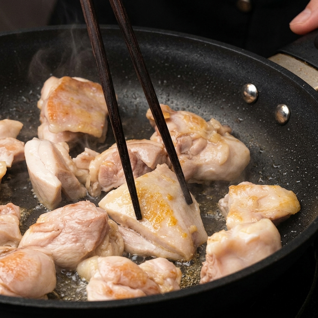フライパンの中で焼き色がついていく鶏肉を、濃い色の木製の箸でひっくり返している様子