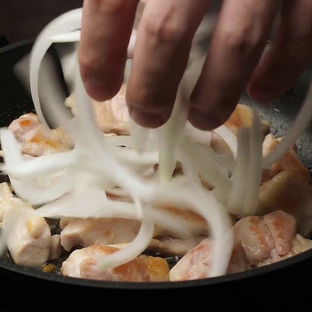フライパンの中で半分ほど火が通った鶏肉の上に、薄切りにした白い玉ねぎをスライスして投入している手元