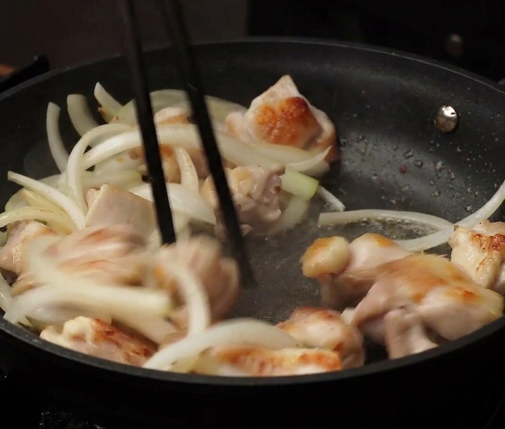 黒いノンスティック加工のフライパンの中で、一口大の鶏肉とスライスした玉ねぎを箸で混ぜ合わせながら炒めている様子