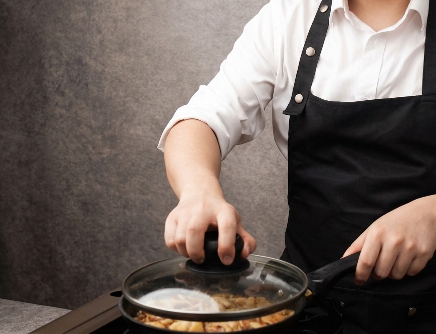 薄茶色のタレで煮込まれている鶏肉と玉ねぎのスライスが入ったフライパンに、透明なガラスの蓋をかぶせている手元