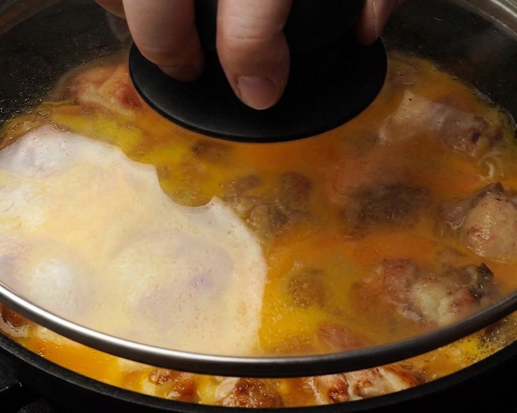 フライパンにかぶせたガラス蓋越しに見える、鶏肉を部分的に覆うように固まりつつある黄色い卵液のクローズアップ