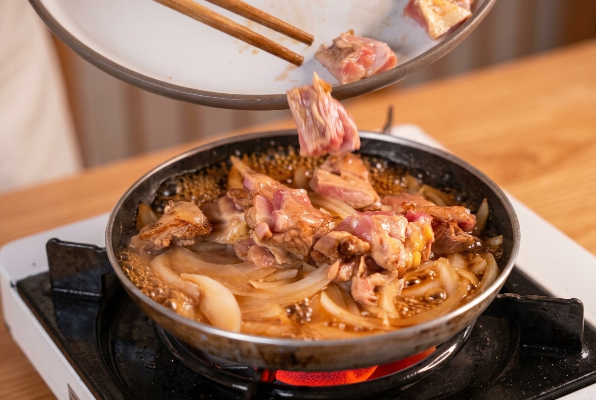 玉ねぎと出汁がグツグツと煮立つフライパンに、箸で香ばしく焼いた鶏肉を戻し入れている様子。