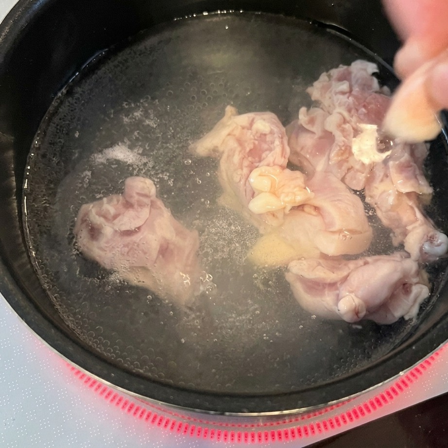 赤色のIHクッキングヒーターの上にある、沸騰したお湯の入った鍋に生の鶏肉を投入する両手。
