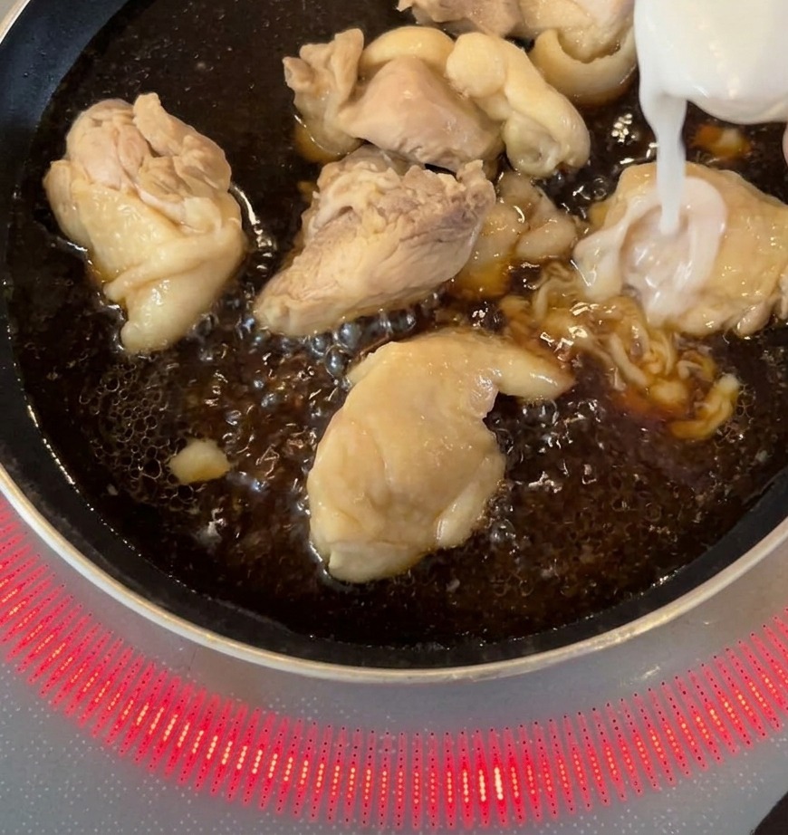 濃いタレで鶏肉が煮込まれているフライパンに、スライスした白い玉ねぎが加えられている様子。