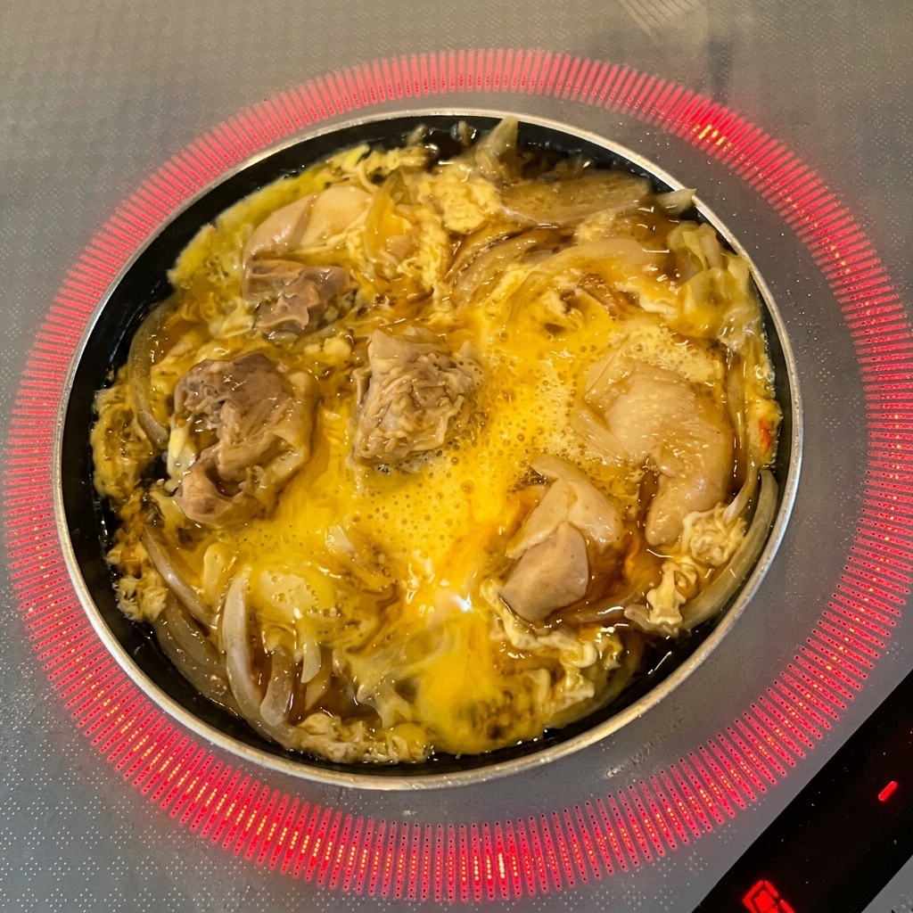 半分ほど火が通った卵と鶏肉の上に、新鮮な緑の三つ葉の葉が散らされている様子。