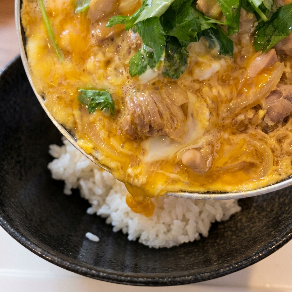温かい白米が入った丼に、フライパンを傾けて調理済みのとろとろの鶏肉と卵の具を滑らせるようにのせている様子。