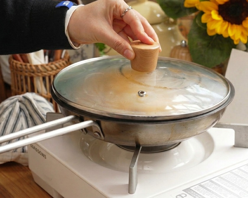 フライパンにガラスの蓋をかぶせ、カツと卵の料理を蒸気で調理している手元。