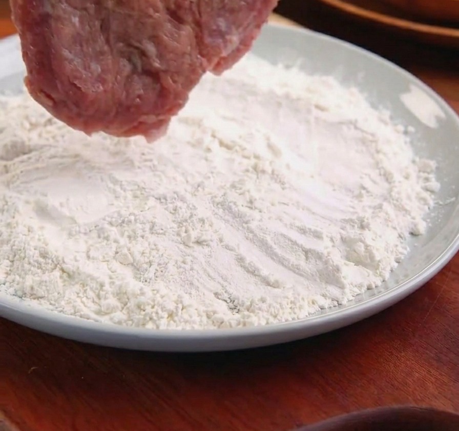 トングで豚肉を持ち上げ、薄力粉の入った皿に入れている様子