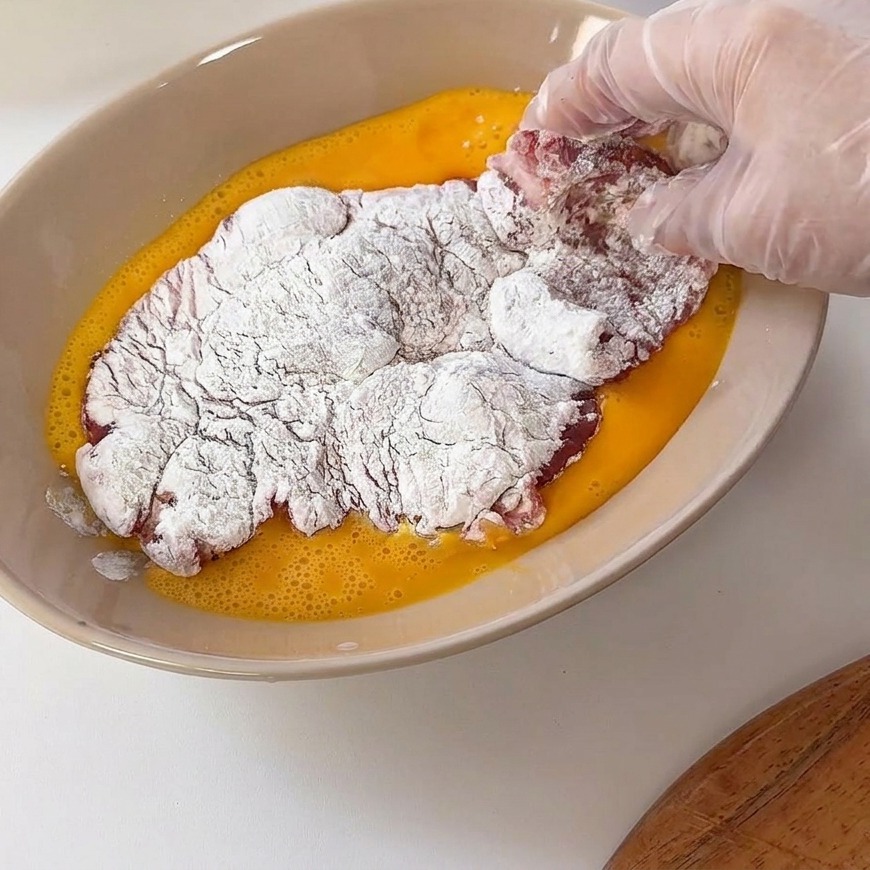薄力粉をまぶした豚肉を、溶き卵の入ったボウルに入れている様子。