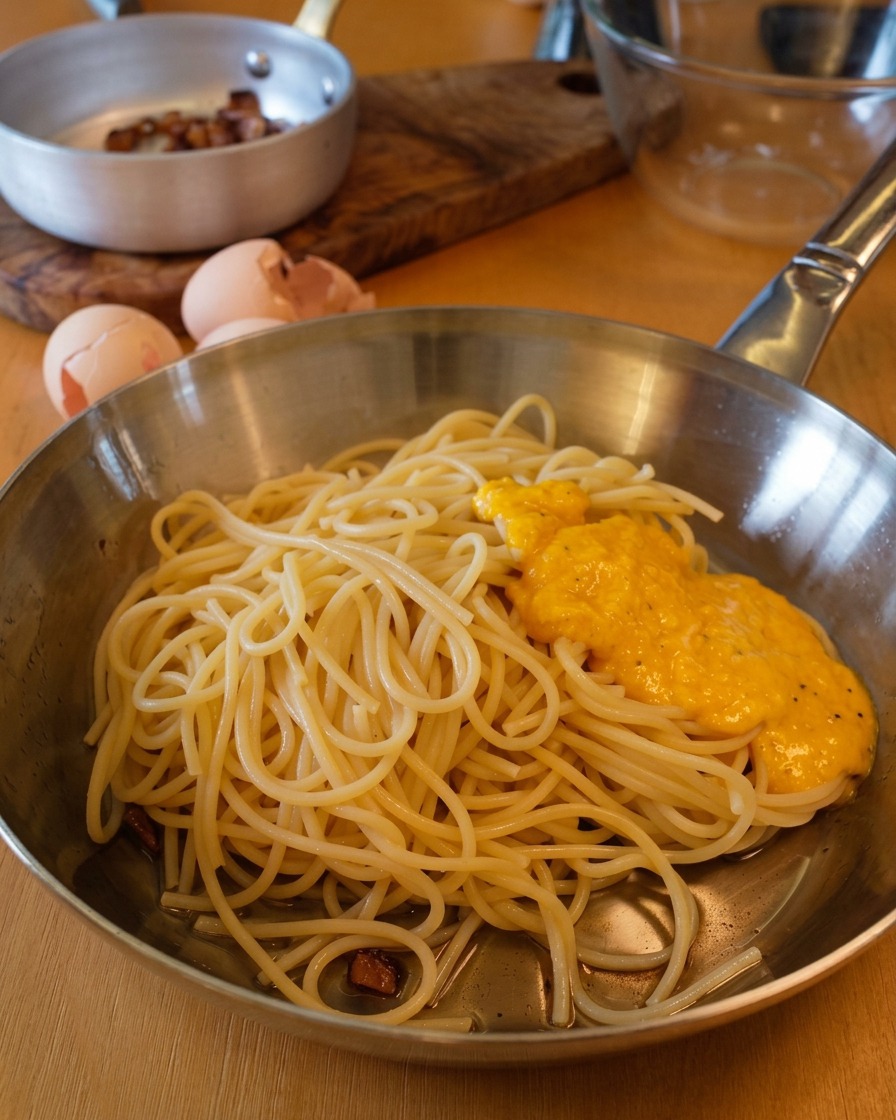 フライパンの中のスパゲッティの上に、黄金色の卵とチーズのソースがかけられている様子。