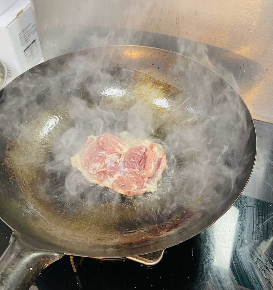 湯気と油が揺らめく熱い中華鍋に、生の鶏もも肉を1枚入れている様子。