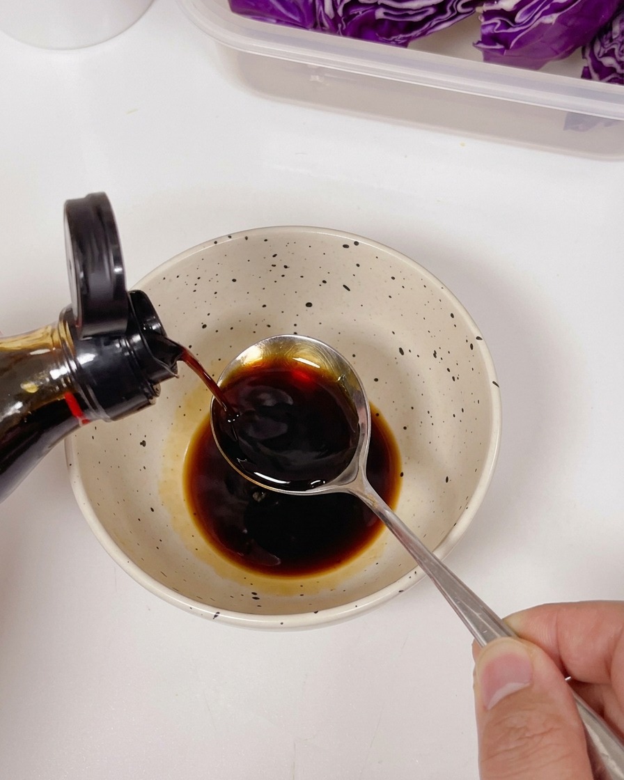 濃い色のソースと調味料が入ったボウルに、ボトルからスプーンを使って薄口醤油を注いでいる様子。