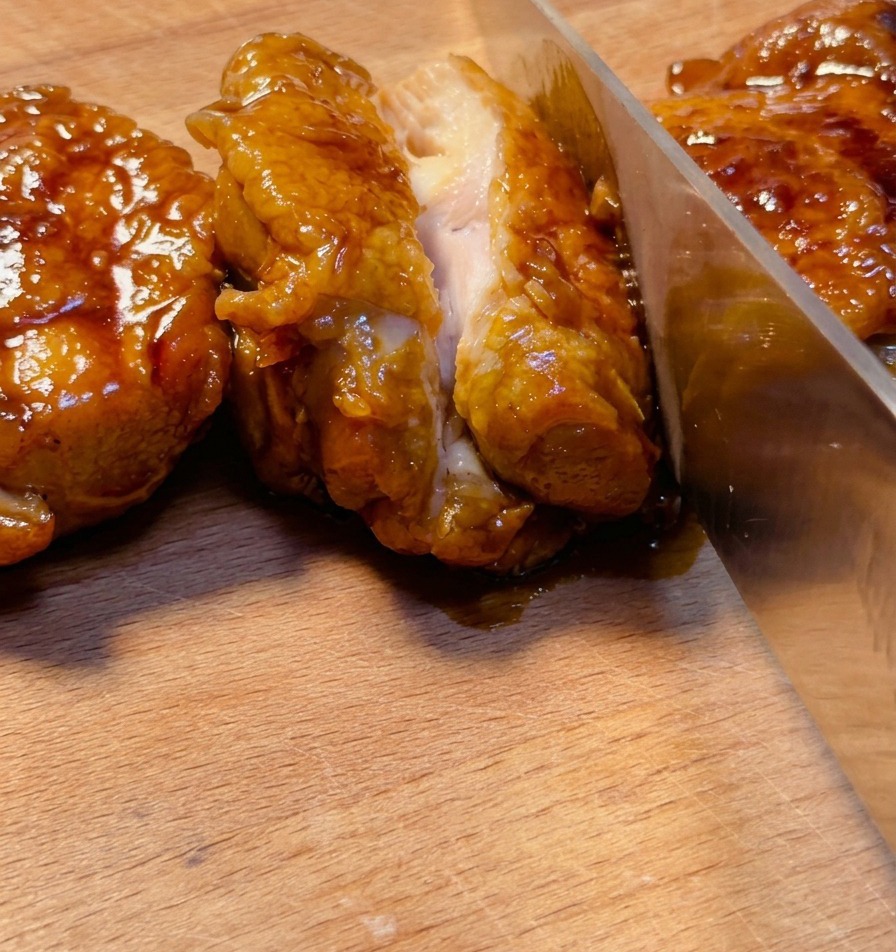 木製のまな板の上で、艶やかなタレが絡んだ照り焼きチキンを鋭い包丁で切り、ジューシーな断面を見せている様子。